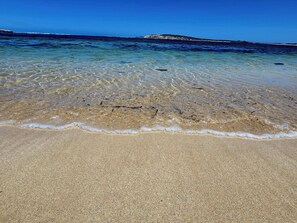 A beach with clear shallow waters, a sandy shore, and a distant rocky outcrop.