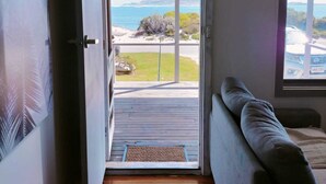 A room with a wooden floor, a sofa, a door leading to a balcony with a view of the sea and a beach.