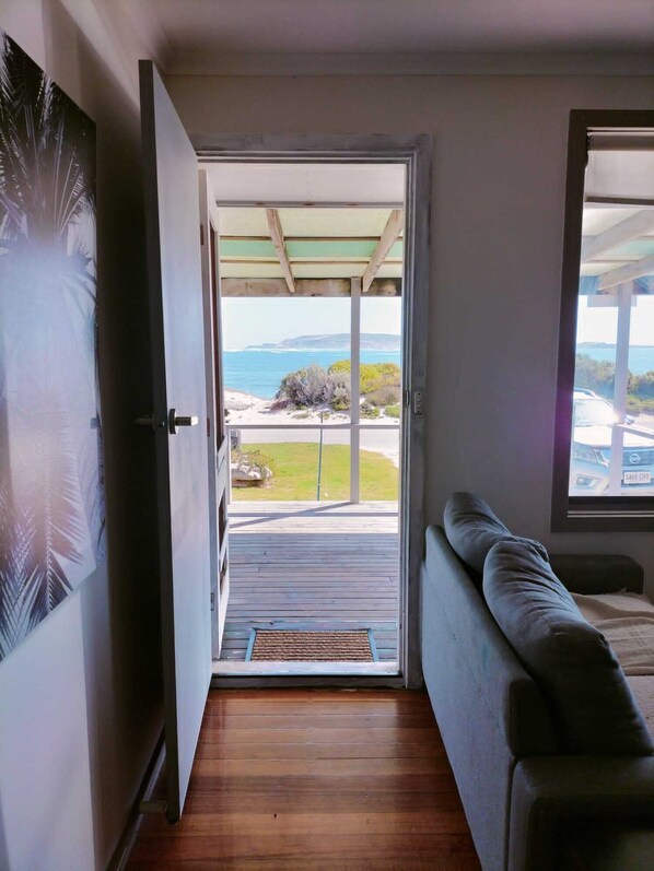 A room with a wooden floor, a sofa, a door leading to a balcony with a view of the sea and a beach.