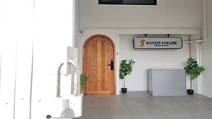 Een receptiegedeelte met een houten deur, een bord waarop 'SHADE HOUSE Bangkok Oldtown' staat, en twee potplanten.
