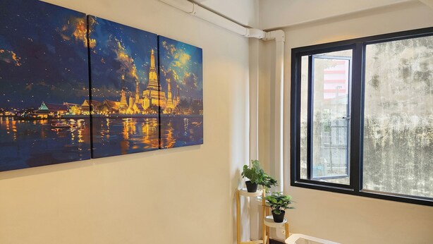 Een kamer met een groot schilderij van een nachtelijk tafereel, een raam met uitzicht op gebouwen, en een kleine plant op een standaard.