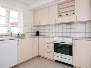 Une cuisine moderne avec des meubles en bois clair, un lave-vaisselle blanc, un four encastré et une fenêtre agrémentée d’une plante posée sur le rebord.