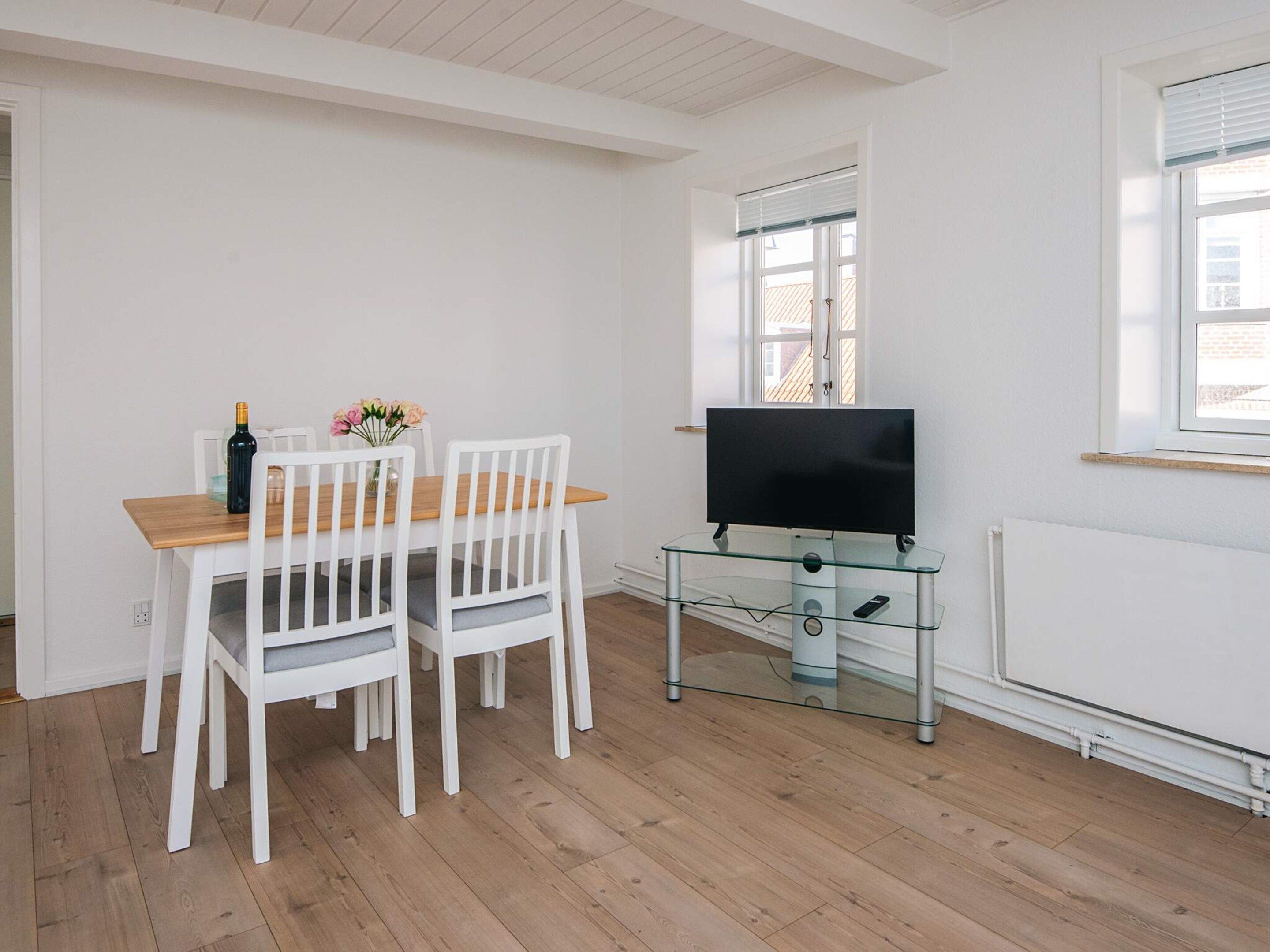 A room with a wooden dining table, white chairs, a glass TV stand, and a window with blinds.