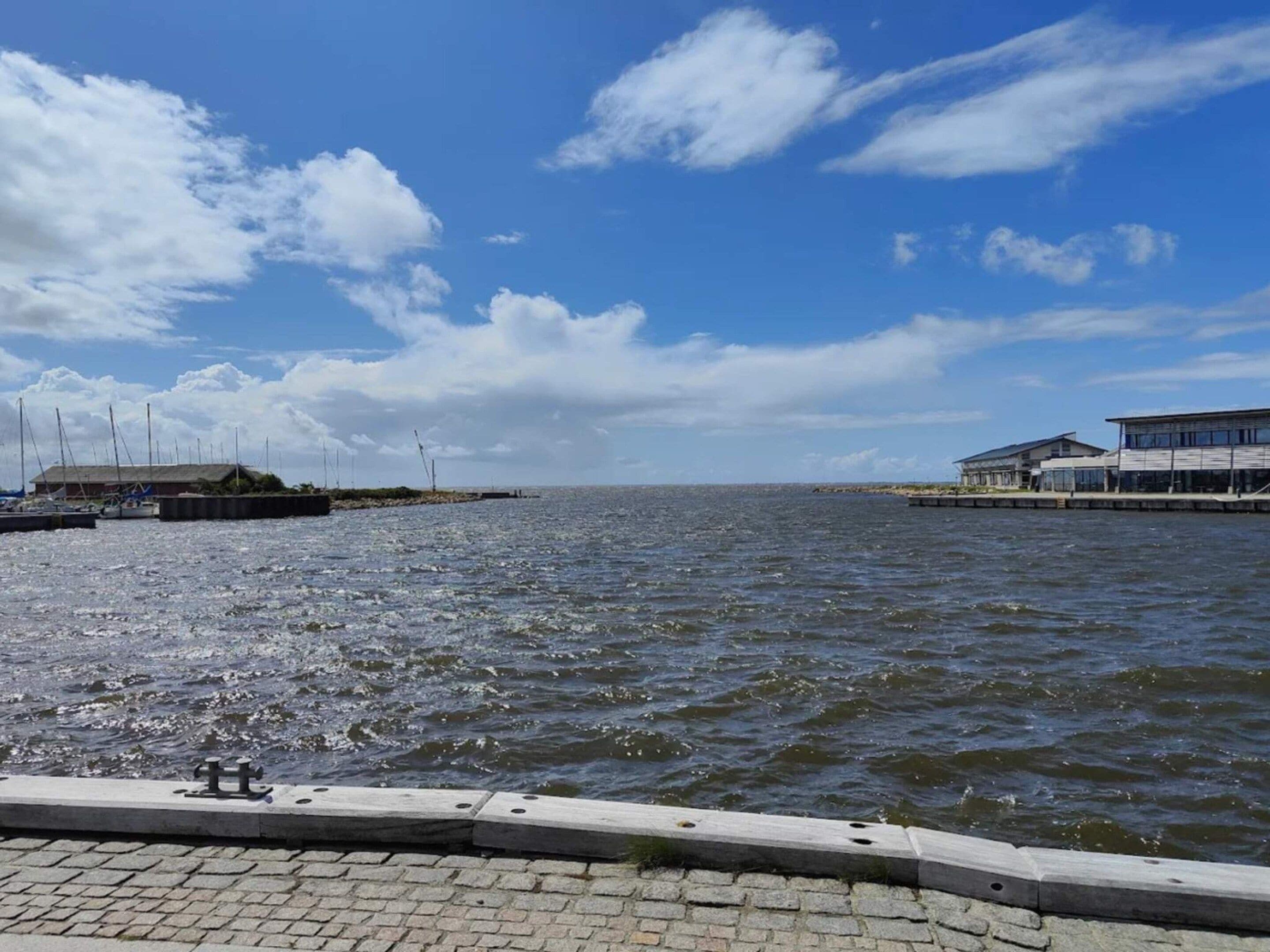 A waterfront view with a paved walkway, boats, and buildings along the shore.