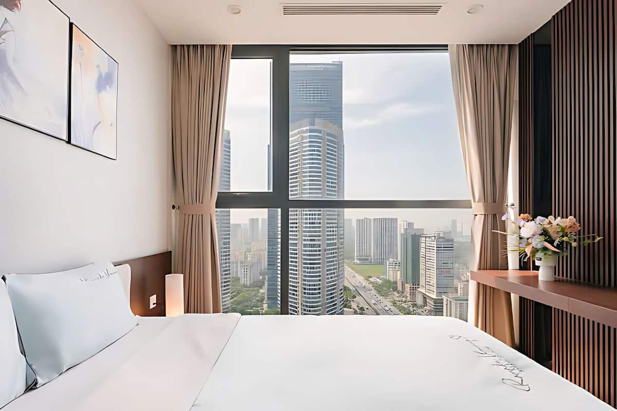 A modern hotel room with a large bed, a view of the cityscape, and a vase of flowers on the nightstand.