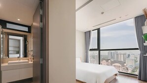 Een moderne hotelkamer met een groot bed, een badkamer met een wastafel en spiegel, en uitzicht op de stad door grote ramen.