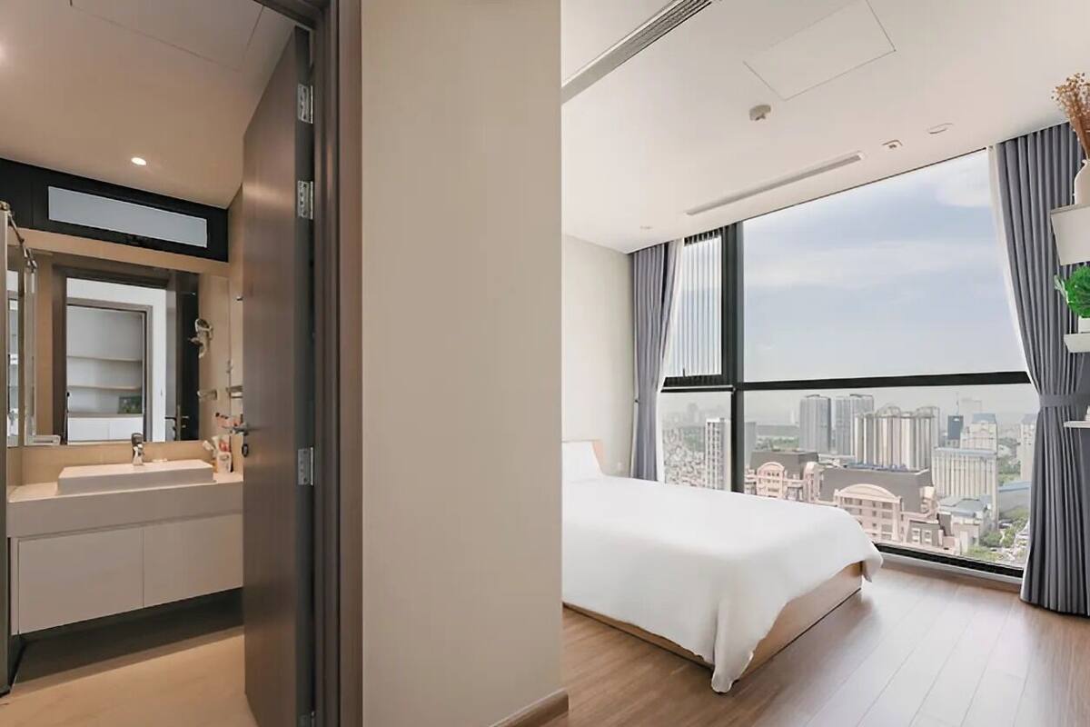 A modern hotel room with a large bed, a bathroom with a sink and mirror, and a city view through large windows.