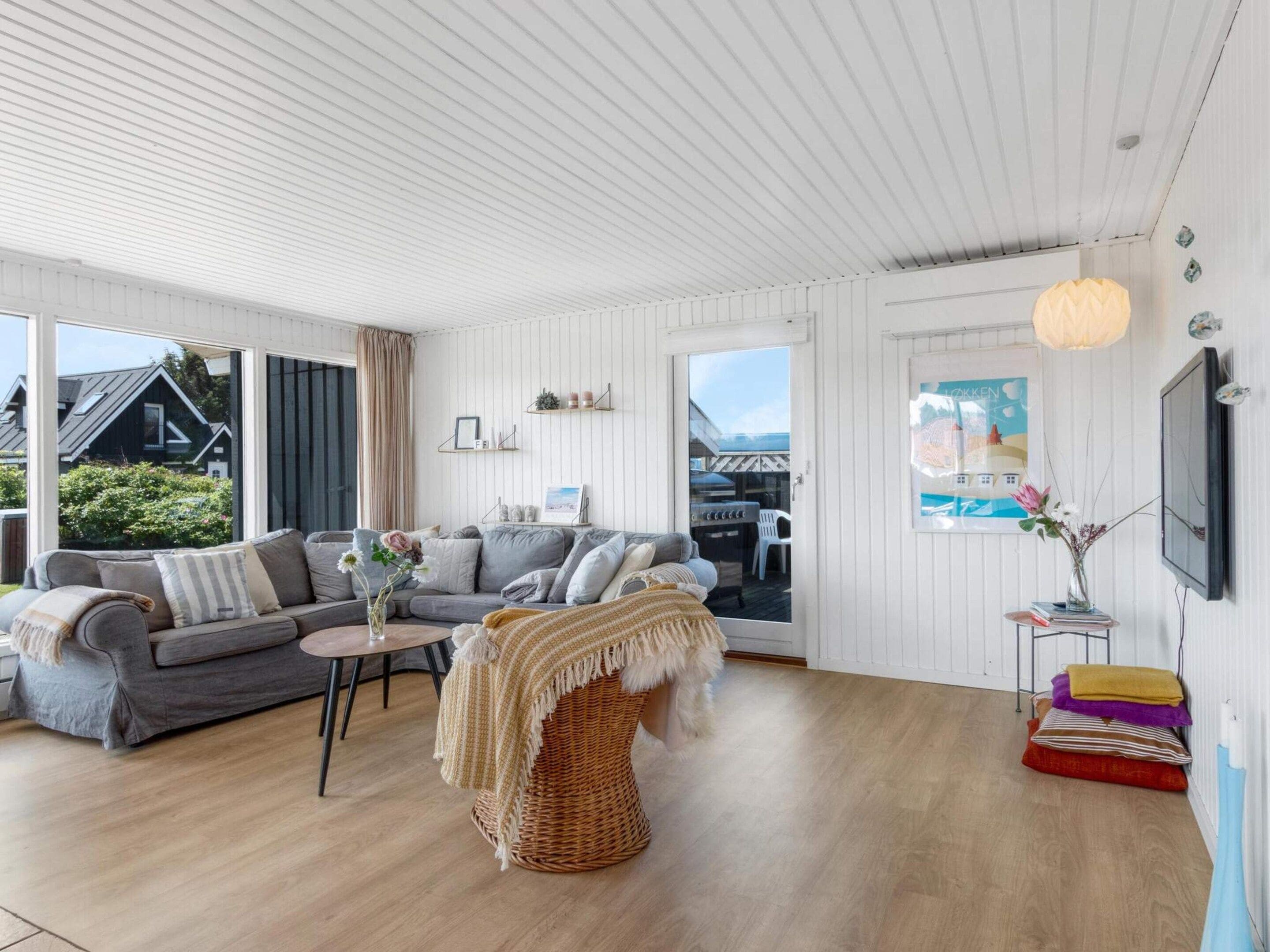 Una sala de estar moderna con un sofá, una mesa redonda y una silla de mimbre. Hay un televisor montado en la pared y una ventana con vistas al exterior.