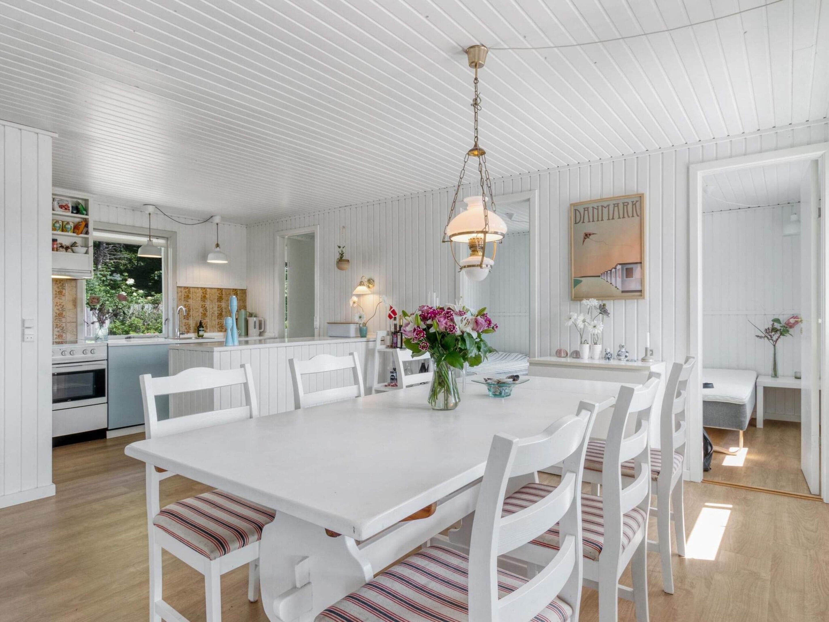 A white dining area with a table and chairs, a hanging light fixture, and a kitchen in the background.