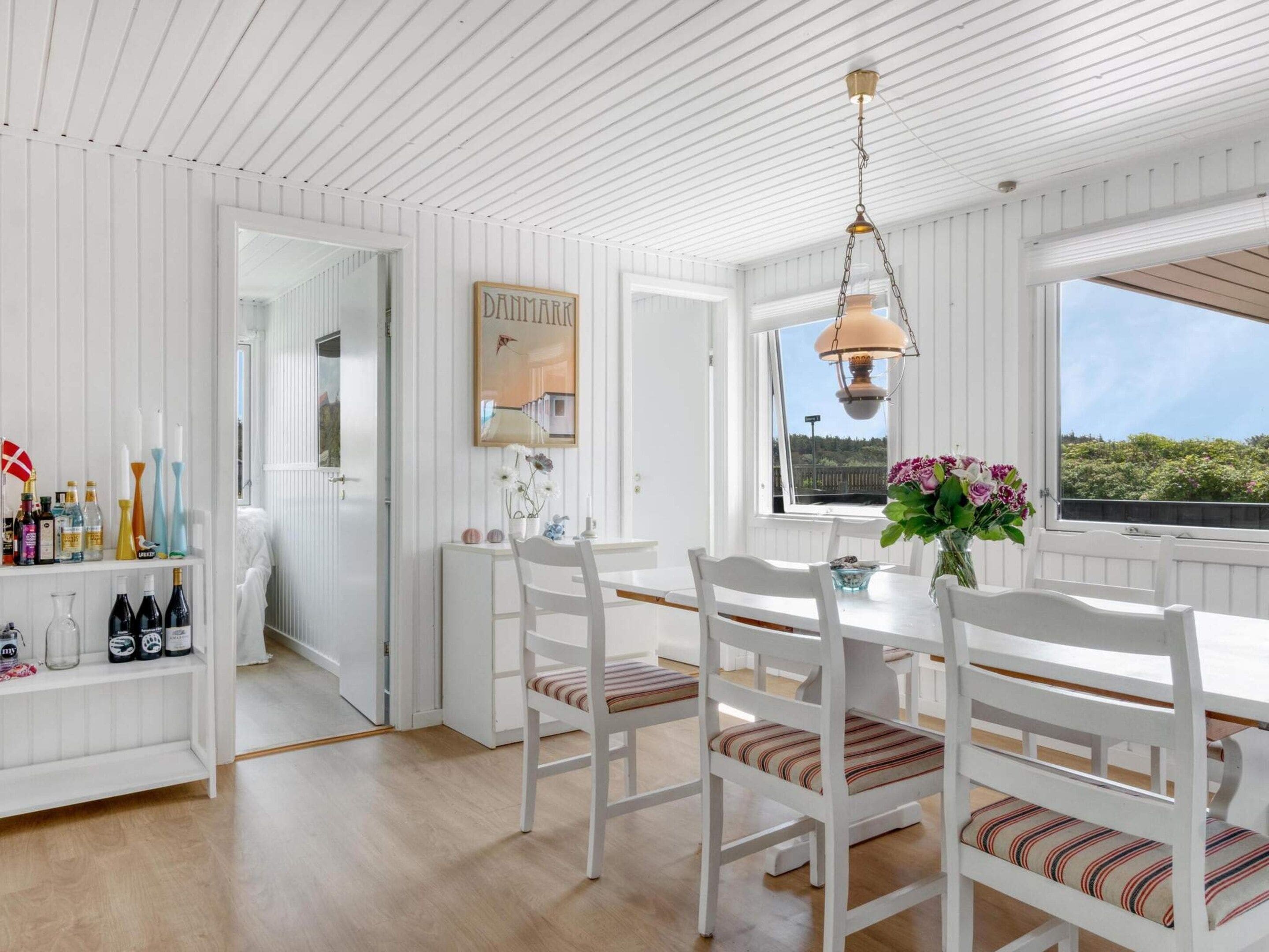 Un coin repas avec des meubles en bois peints en blanc, un luminaire suspendu et une vue par la fenêtre.