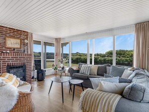 A spacious living room with a brick fireplace, a grey sofa set, a wicker chair, and a large window offering a view of greenery.