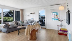 Uma sala de estar ampla com um sofá cinzento, uma mesa de centro redonda e uma poltrona de vime. Há uma TV montada na parede e uma janela com vista para as casas lá fora.
