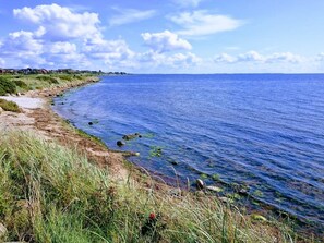 A coastal landscape with a sandy beach, clear blue water, and distant buildings. - 2 Person Holiday Home in Ringkobing (Ringkobing)