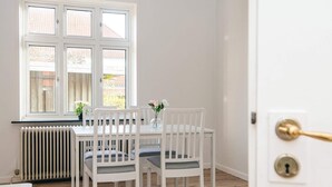 A dining area with a white table and chairs, a window with a view of a garden, and a radiator under the window. - 2 Person Holiday Home in Ringkobing (Ringkobing)