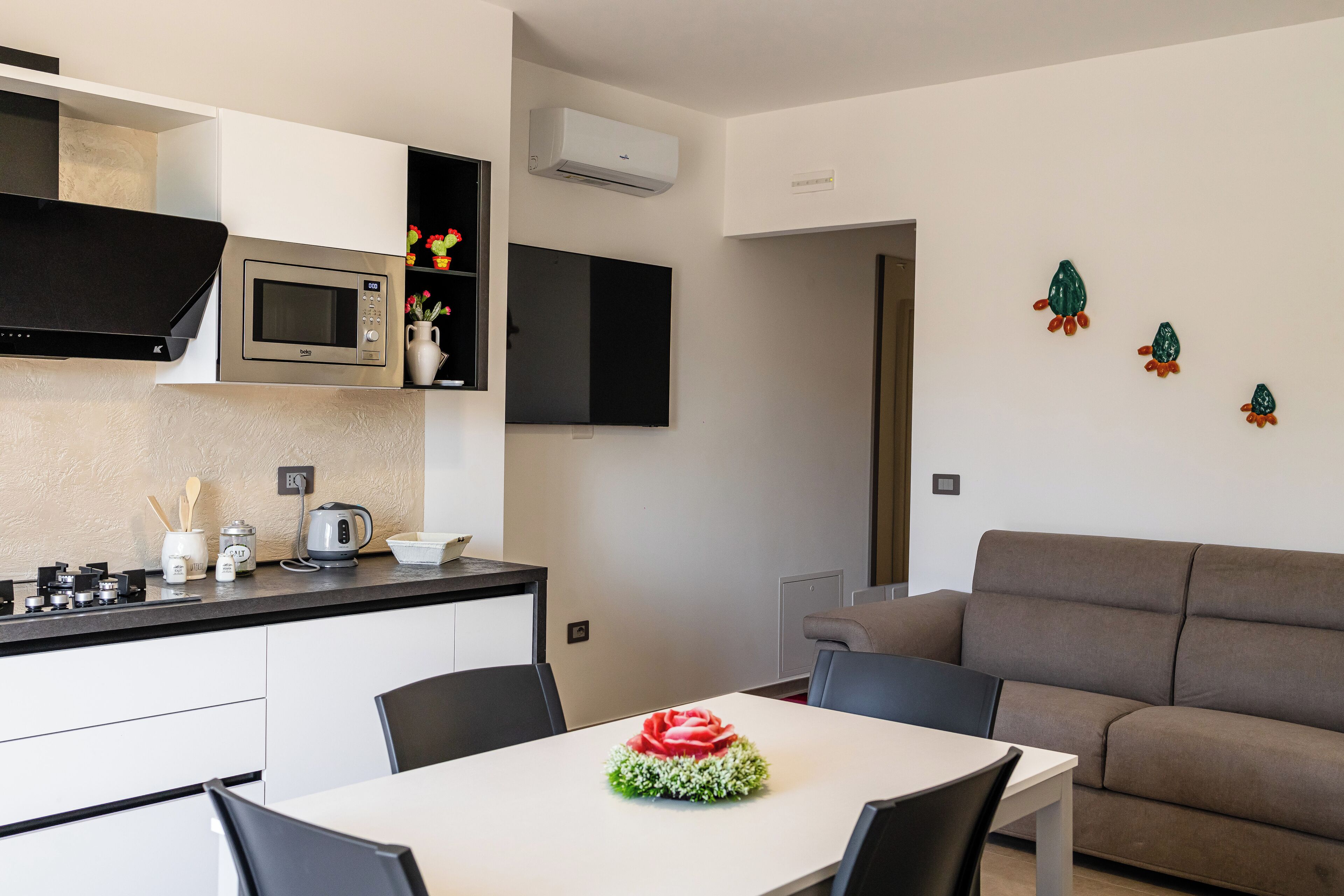 A modern kitchen with a built-in microwave, a kettle, and a coffee maker. There is a dining table with chairs and a decorative rose centerpiece. A brown sofa is visible in the background.