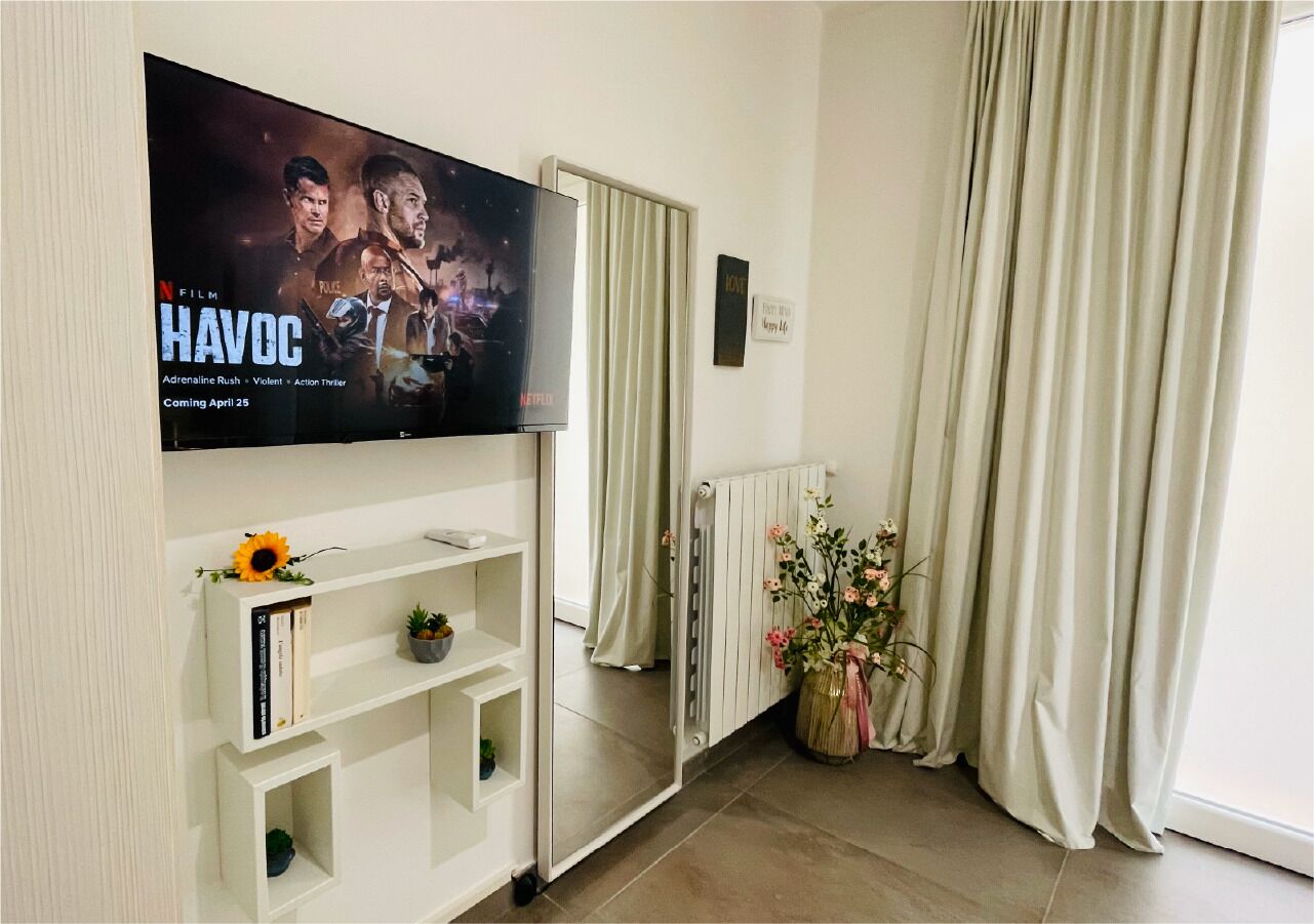 Un salon avec un téléviseur à écran plat fixé au mur, un meuble de rangement blanc contenant des livres et agrémenté d’une décoration en forme de tournesol, un radiateur et un vase de fleurs.