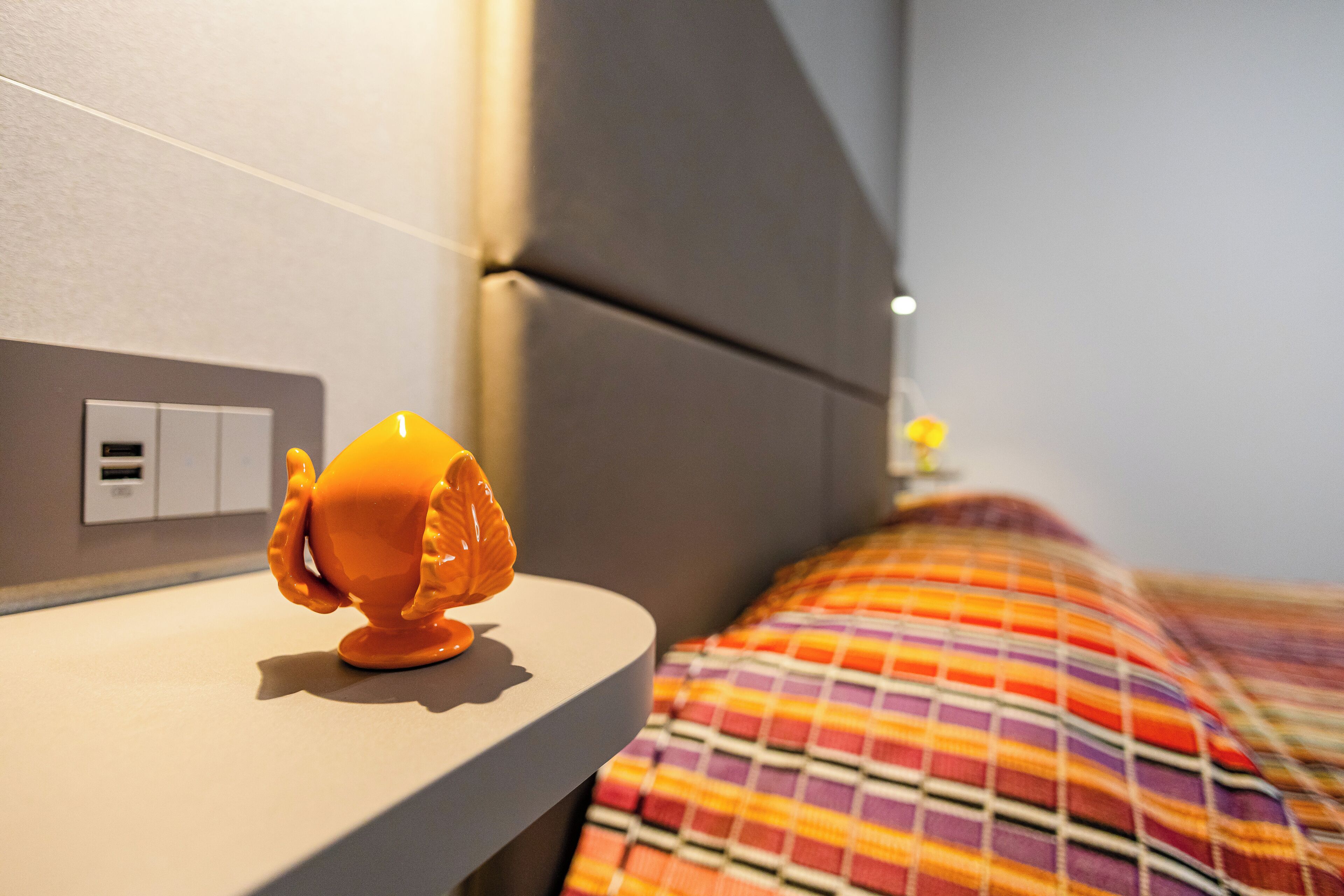 Una cama con una colcha a rayas de colores, una mesita de noche con un objeto naranja pequeño y un enchufe de pared.