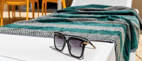 A sun lounger with a pair of sunglasses, a patterned blanket, and a white chair in the background.