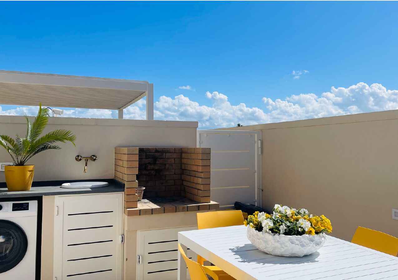 Una cocina exterior compacta con horno de ladrillo, lavadora y comedor con sillas amarillas y mesa blanca.