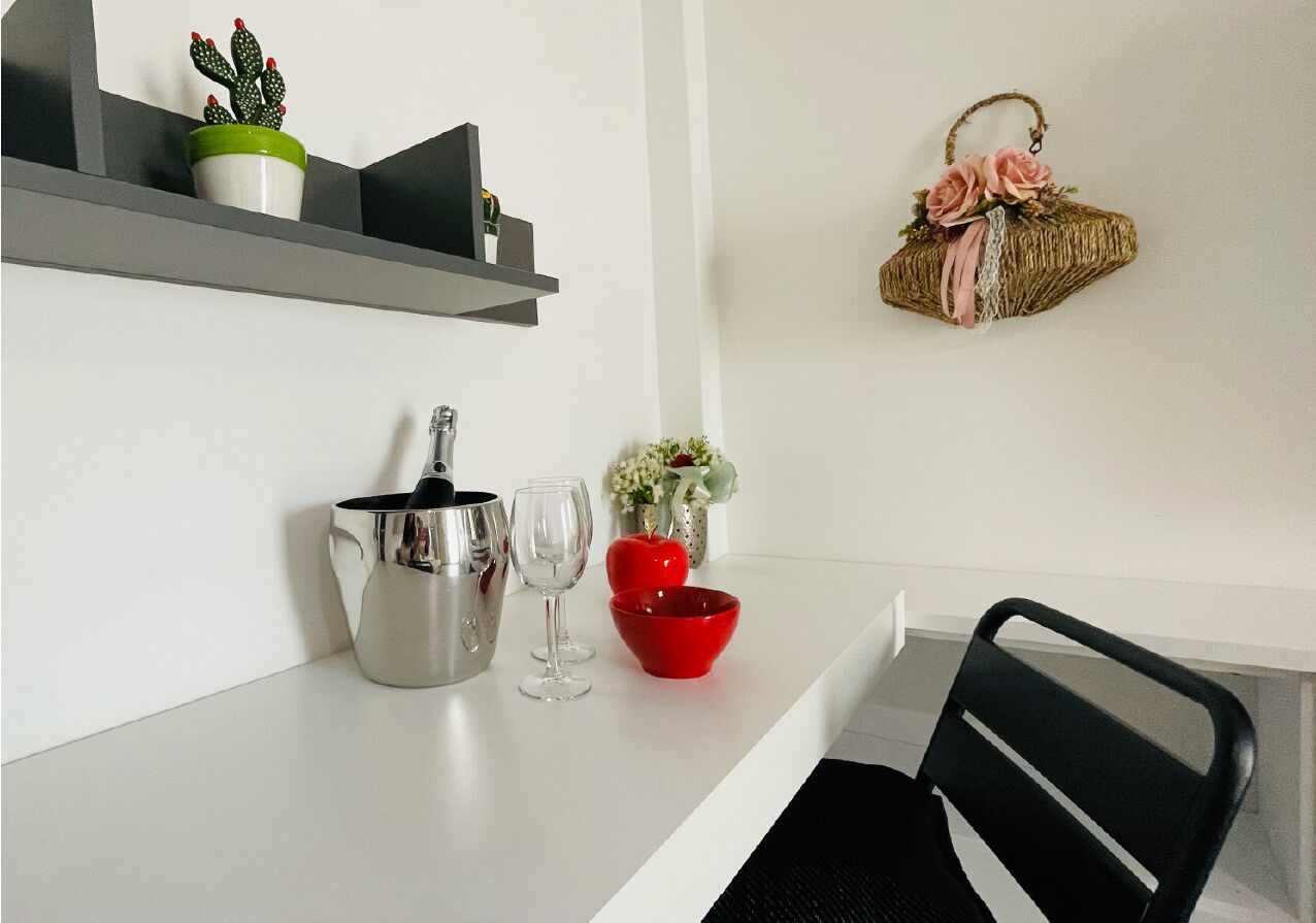 A kitchen counter with a kettle, wine glasses, and a red bowl. There is a potted cactus on a shelf and a wicker basket with flowers on the wall.
