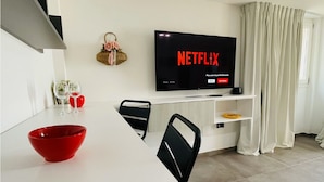 A modern living room with a flat-screen TV displaying the Netflix logo, a white dining table with a red bowl, and a black chair. - La Collina di Montegrappa - Suites with Heated Private Pool - Suites with Shared Pool (Tuglie)