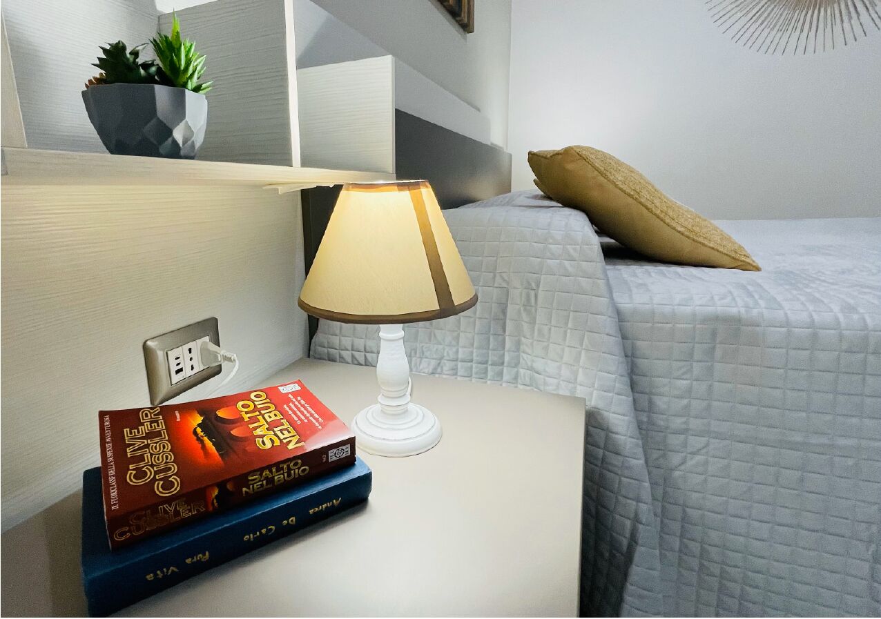 A neatly made bed with a quilted cover, a bedside lamp, and a stack of books on a nightstand.