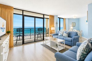 Ein geräumiges Wohnzimmer mit einer großen Schiebeglastür, die auf einen Balkon mit Meerblick führt. Es gibt ein blaues Sofa, einen Couchtisch und einen Fernseher auf einem Lowboard.