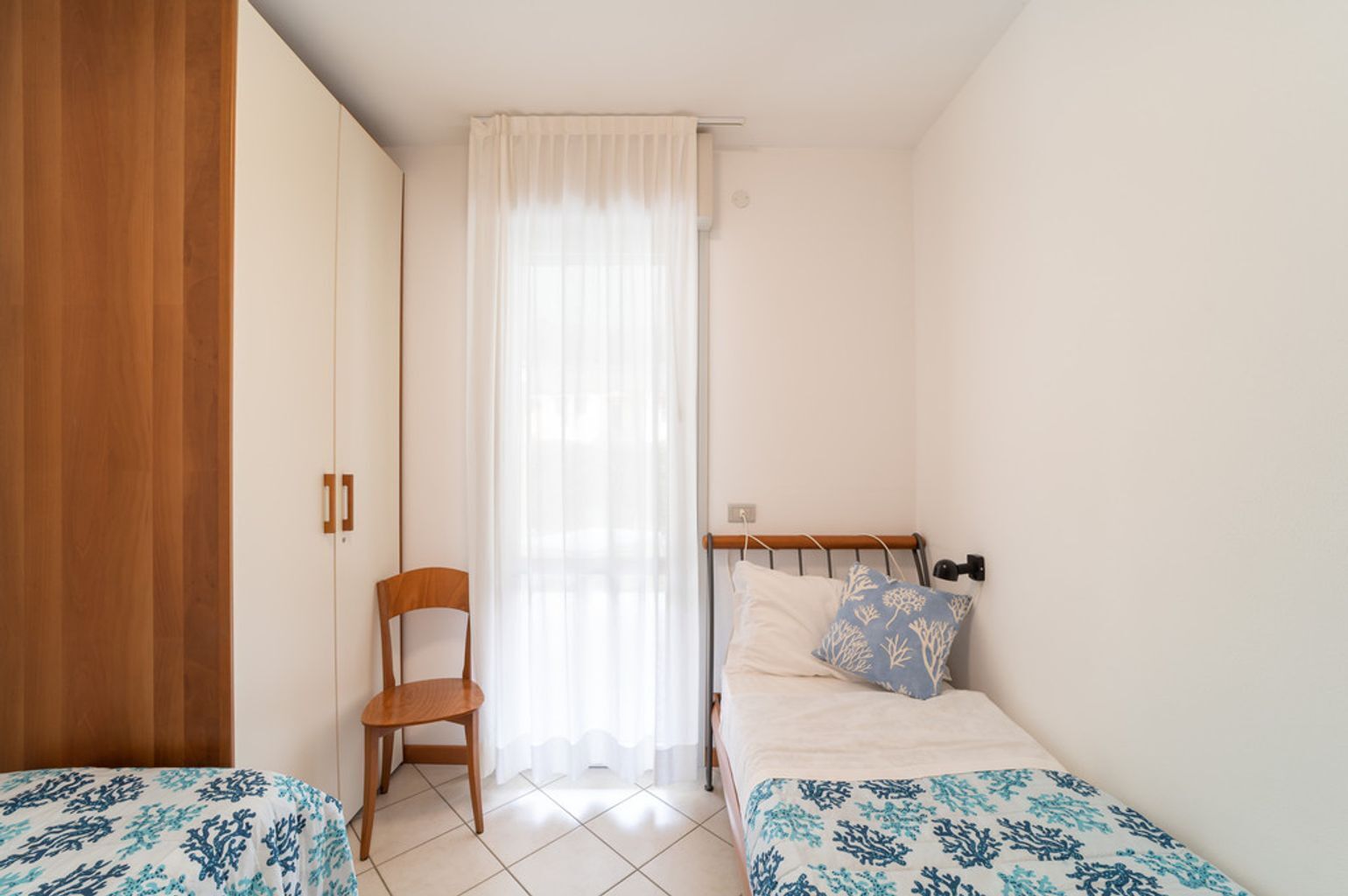 A small, well-lit bedroom with two beds, a wooden wardrobe, a chair, and a window with sheer curtains.