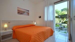 Ein Schlafzimmer mit einem großen Bett, einem Nachttisch, einer Lampe und Blick auf einen Garten durch eine Schiebetür.
