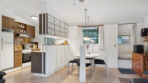 Une cuisine moderne avec des meubles blancs, un coin repas avec une table blanche et des chaises noires, et un salon avec un canapé et un téléviseur.