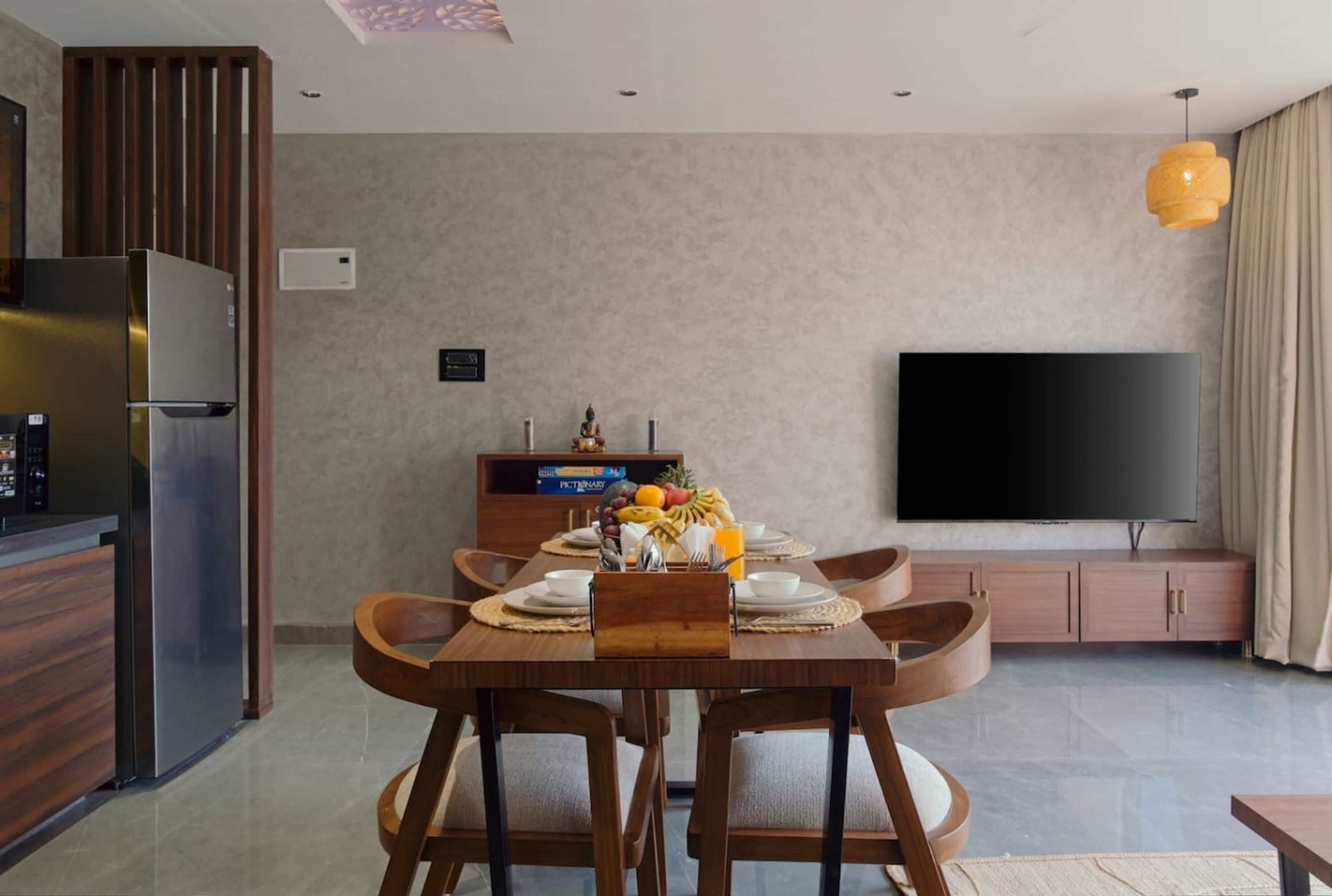 A modern living room with a dining table, wooden chairs, a flat-screen TV, a kitchen area with a refrigerator, and a wooden cabinet.