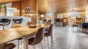 Un intérieur de chalet chaleureux avec un plafond en bois, un coin repas avec une longue table et des chaises, une cuisine équipée d’appareils modernes et un salon avec des fauteuils en cuir et un téléviseur.