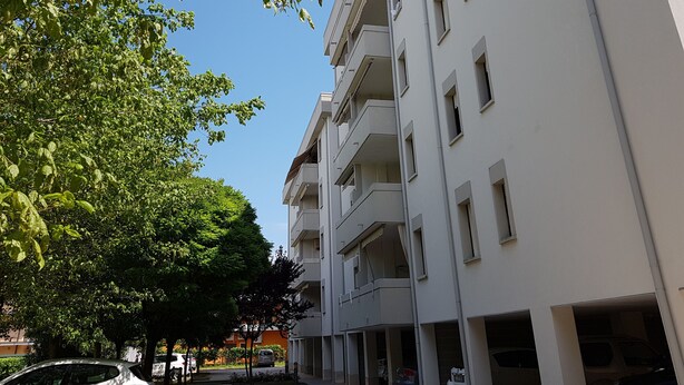 Un immeuble résidentiel à plusieurs étages, doté de balcons, d’un garage et bordé d’arbres au premier plan.