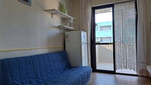 Une pièce avec un canapé bleu, un réfrigérateur blanc, une petite étagère sur laquelle se trouve un vase vert, et une porte-fenêtre coulissante donnant sur un espace extérieur.