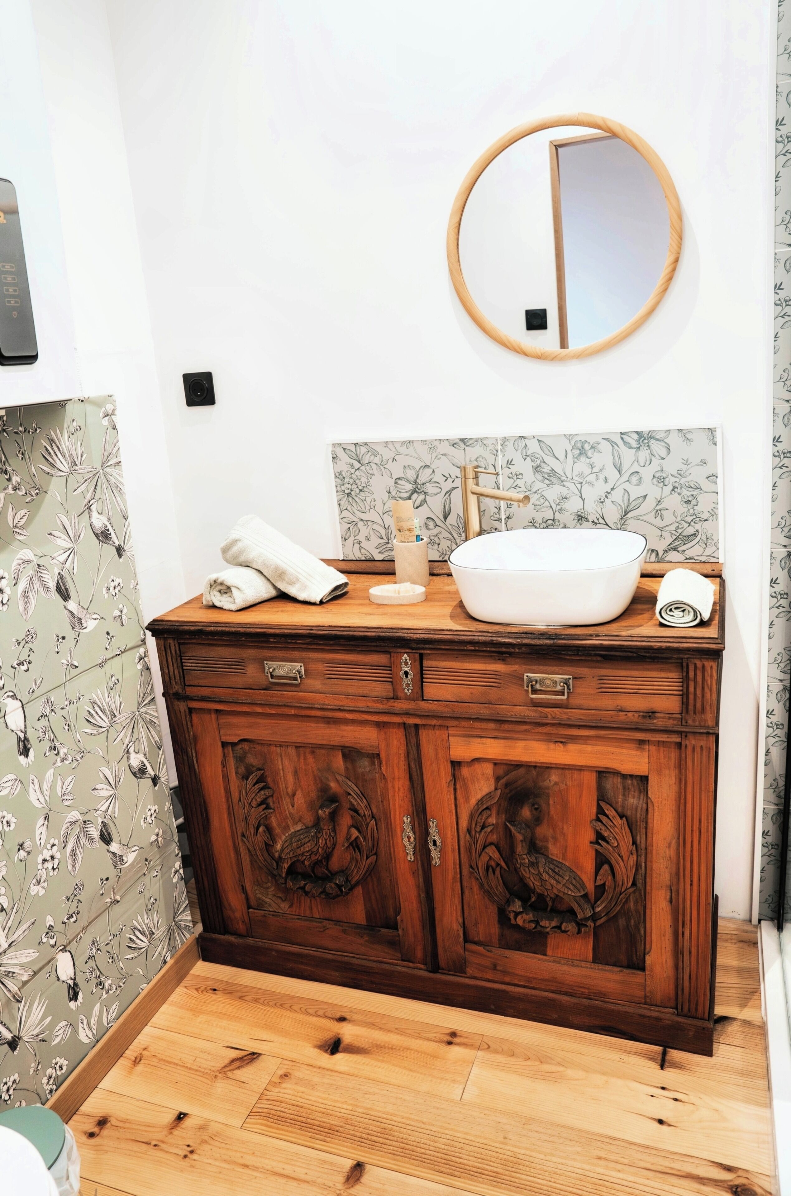 Une coiffeuse en bois avec un lavabo blanc, un miroir rond et un papier peint à motifs floraux.