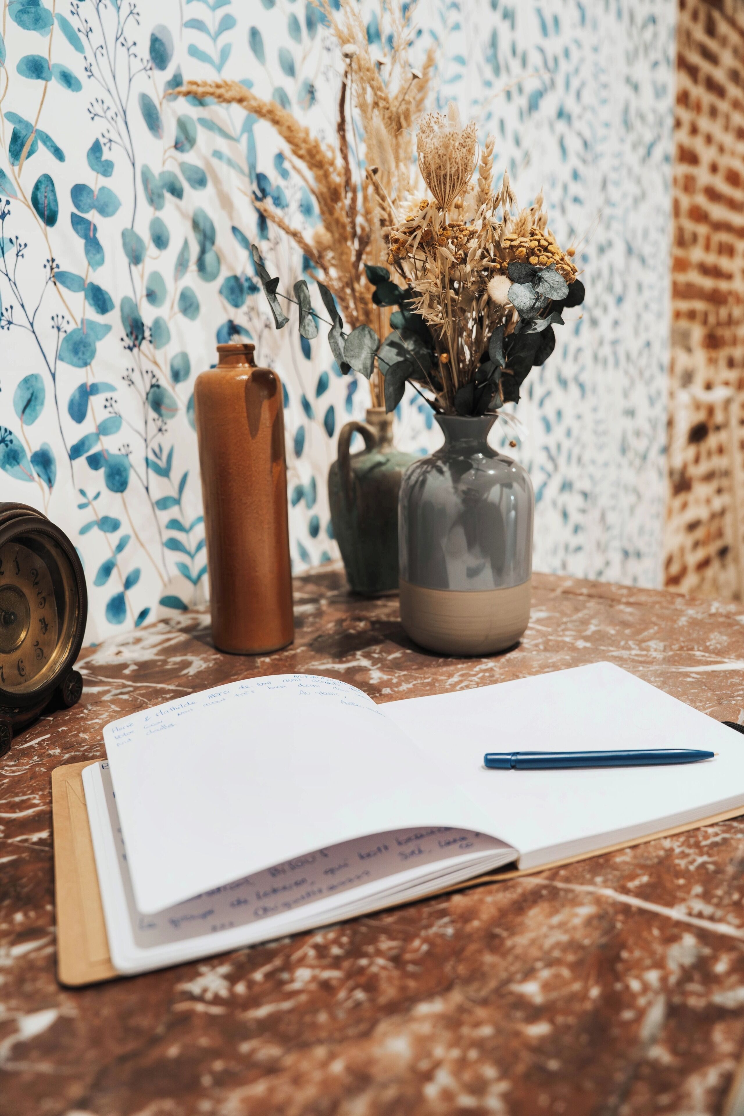 Uma mesa com um caderno, uma caneta e dois vasos com plantas secas.