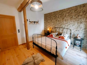 Une chambre avec une porte en bois, un lit avec un cadre en métal, un mur en pierre, un luminaire suspendu et une petite table avec une lampe.