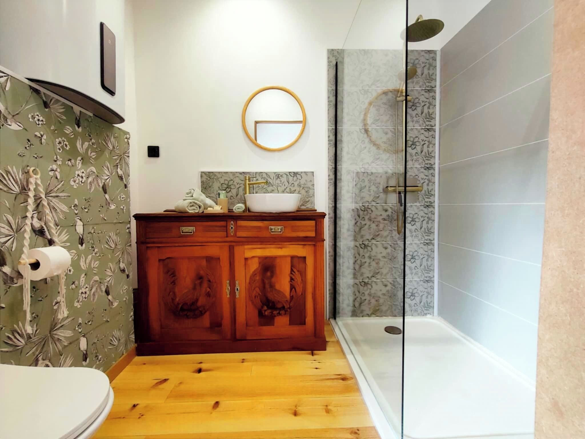 Une salle de bain avec un meuble-lavabo en bois, des toilettes, une douche avec une porte en verre et un papier peint à motifs floraux.