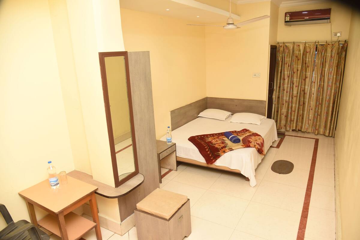 A hotel room with a bed, a desk, a chair, a mirror, a ceiling fan, and an air conditioning unit.