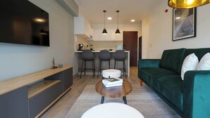 A modern living room with a green sofa, a wooden coffee table, and a flat-screen TV mounted on the wall. There are three bar stools and a kitchen area in the background. - w *| Grand 1BR with Great Balcony in San Isidro (San Isidro)