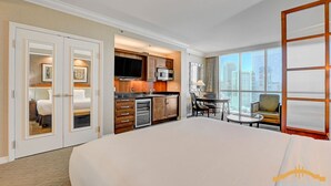 A hotel room with a large bed, a kitchenette, a dining area with a table and chairs, and a city view through large windows.