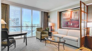 A hotel room with a large window, a sofa, a chair, a coffee table, and a desk with a lamp.