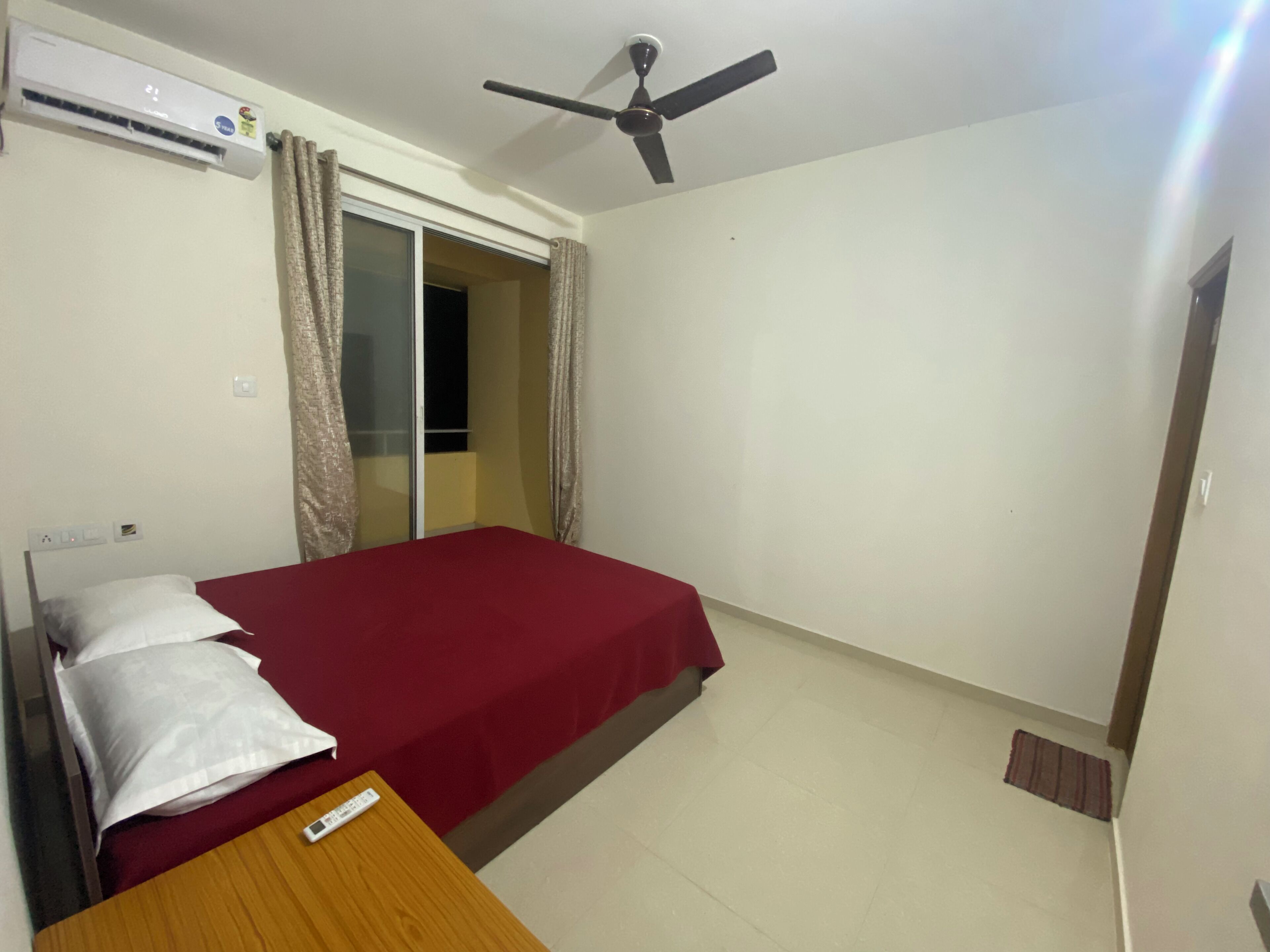 A bedroom with a bed, a wooden table, a ceiling fan, an air conditioning unit, and a sliding door to a balcony.