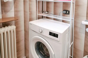 A front-loading washing machine in a bathroom with beige tiles, a white shelf above it, and a white radiator to the left.