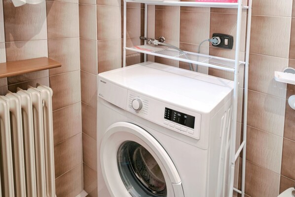 Máquina de lavar frontal numa casa de banho com azulejos bege, uma estante branca acima dela e um radiador branco à esquerda.