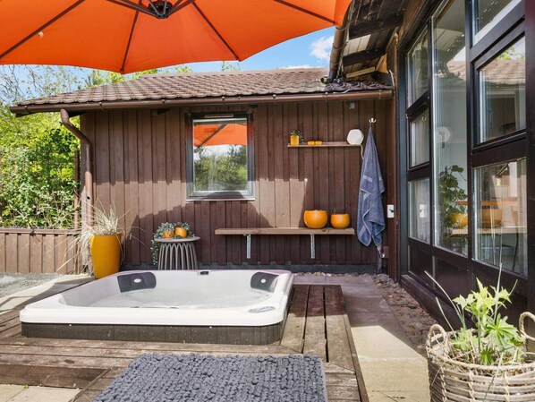 A wooden outdoor hot tub area with a large orange umbrella, a wooden deck, and a window overlooking a garden. - Beautiful Holiday Home in Blokhus (Blokhus)