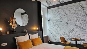 Een moderne hotelkamer met een groot bed, een ronde spiegel, een wand met een bladmotief, een klein rond tafeltje en een stoel.
