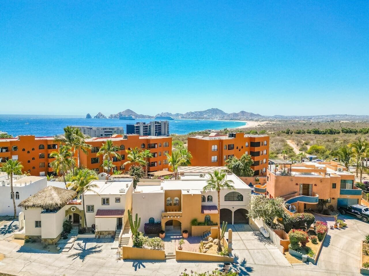 A coastal hotel with multiple buildings, palm trees, and a view of the ocean and mountains.