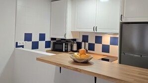 Une cuisine moderne avec des meubles blancs, un micro-ondes noir, un réfrigérateur en acier inoxydable et un plan de travail en bois sur lequel est posé un panier de fruits.
