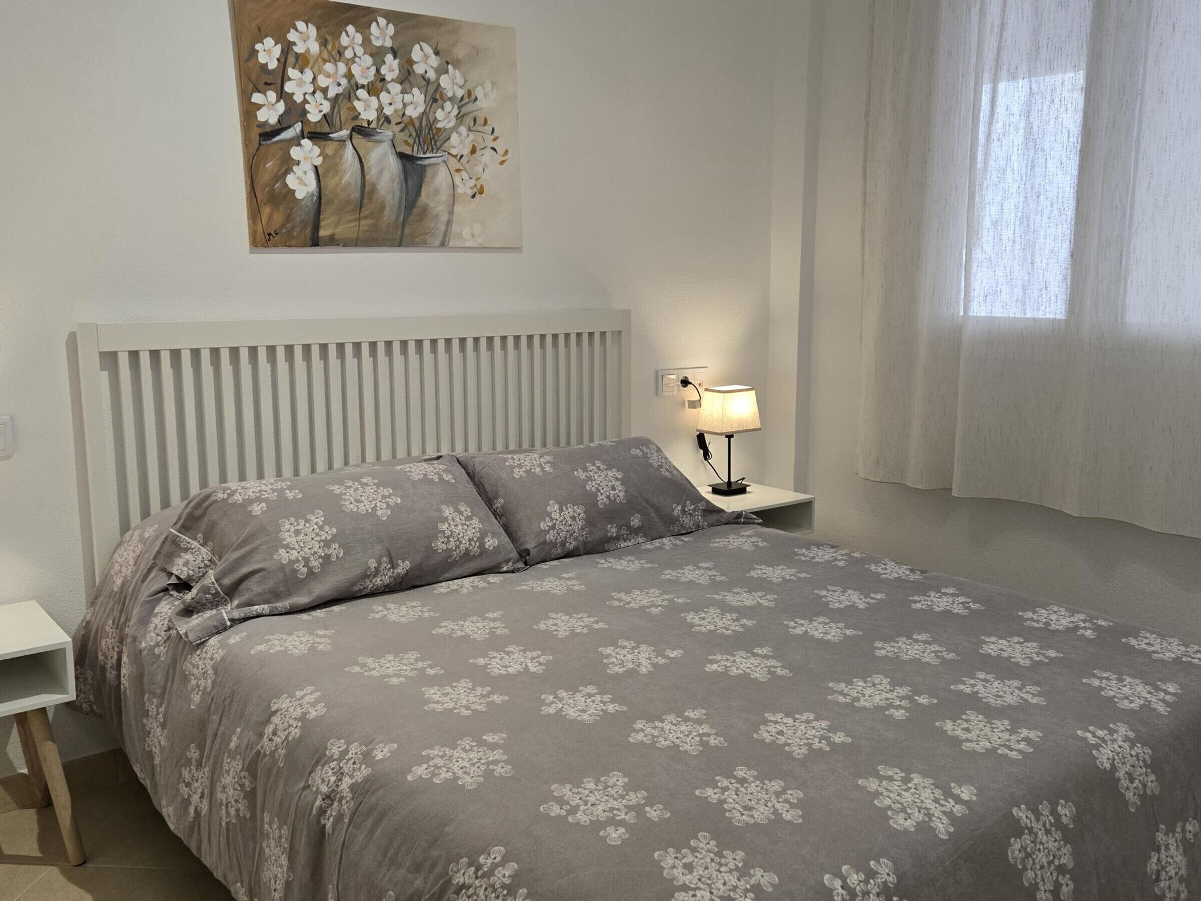 Et soveværelse med en seng, et natbord, en lampe, et maleri og et vindue med gardiner.